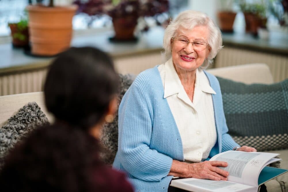 Smiling elderly woman with glasses holding an open book, sitting on a sofa, talking to another person. - Home Instead