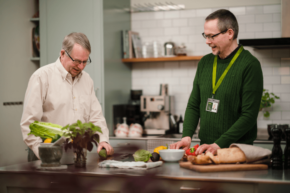 Two men preparing food together in a modern kitchen with fresh vegetables and bread on the worktop. - Home Instead