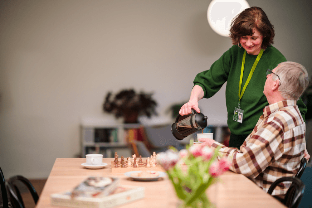Woman in green pours coffee for an older man at a table with chess and flowers. - Home Instead