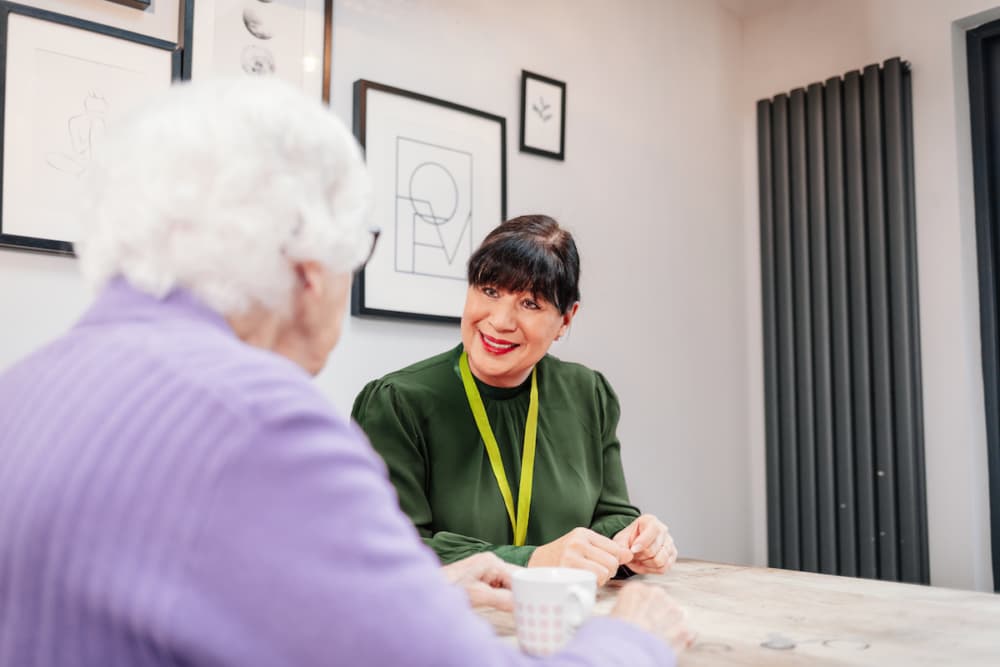 A woman with a lanyard smiles, talking to an older woman in a purple jumper at a table with cups. - Home Instead