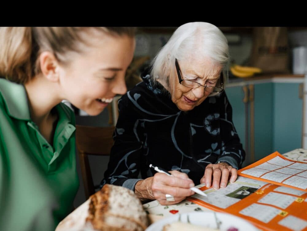 Elderly woman filling in a puzzle book at a table with a smiling young woman beside her. - Home Instead