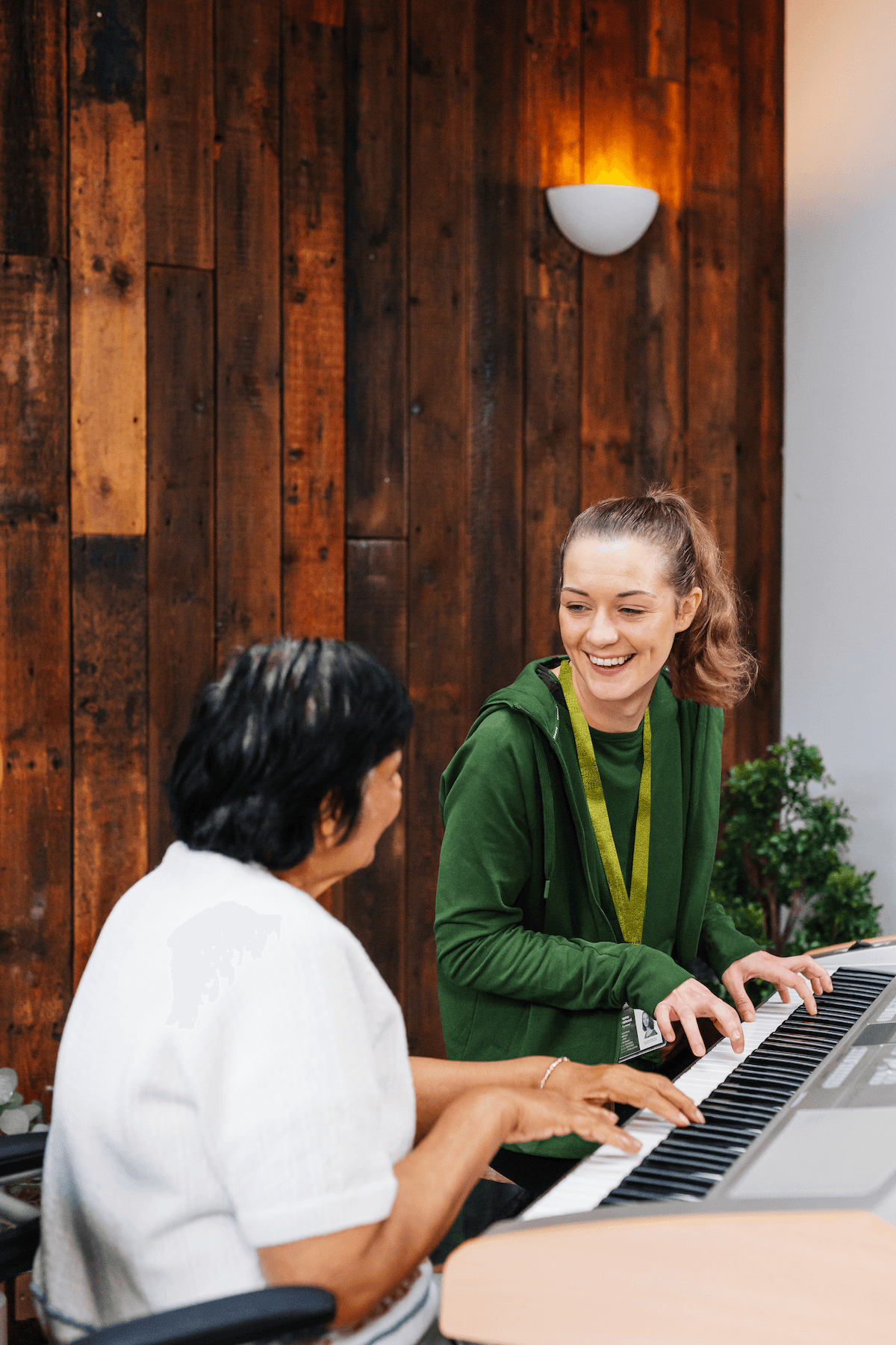 Two women smiling and playing an electric keyboard together in a cosy room with wooden walls. - Home Instead
