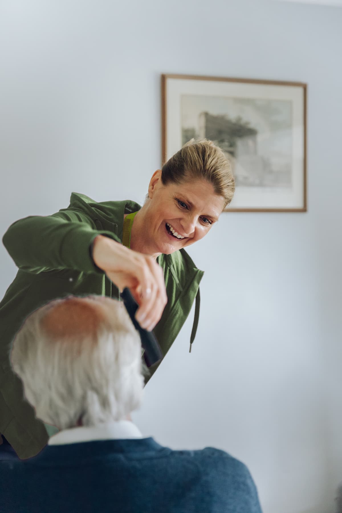 Smiling woman trims an older man's hair indoors, with a framed picture on the wall behind them. - Home Instead