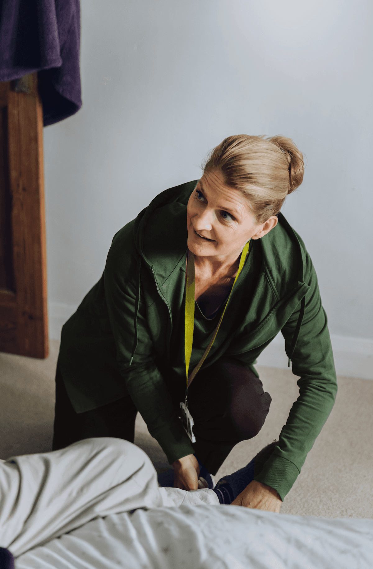 A woman in a green jacket kneels by a bed, helping someone put on socks. - Home Instead
