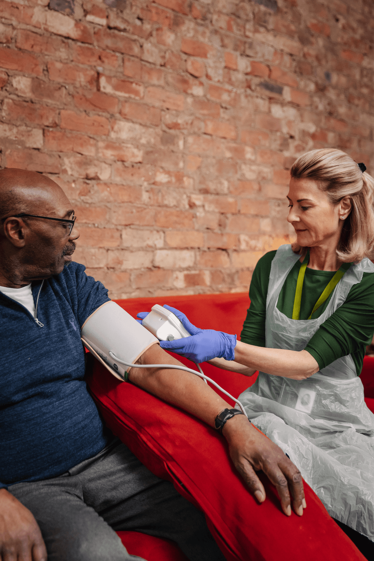A nurse checks a man's blood pressure as they sit on a red sofa against a brick wall. - Home Instead
