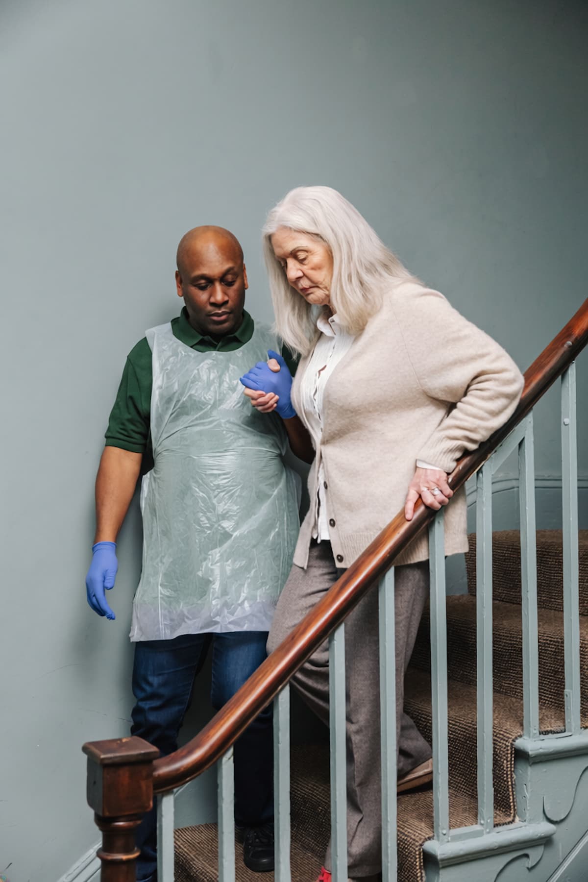 A carer assists an older woman as she carefully descends a staircase, holding her hand for support. - Home Instead