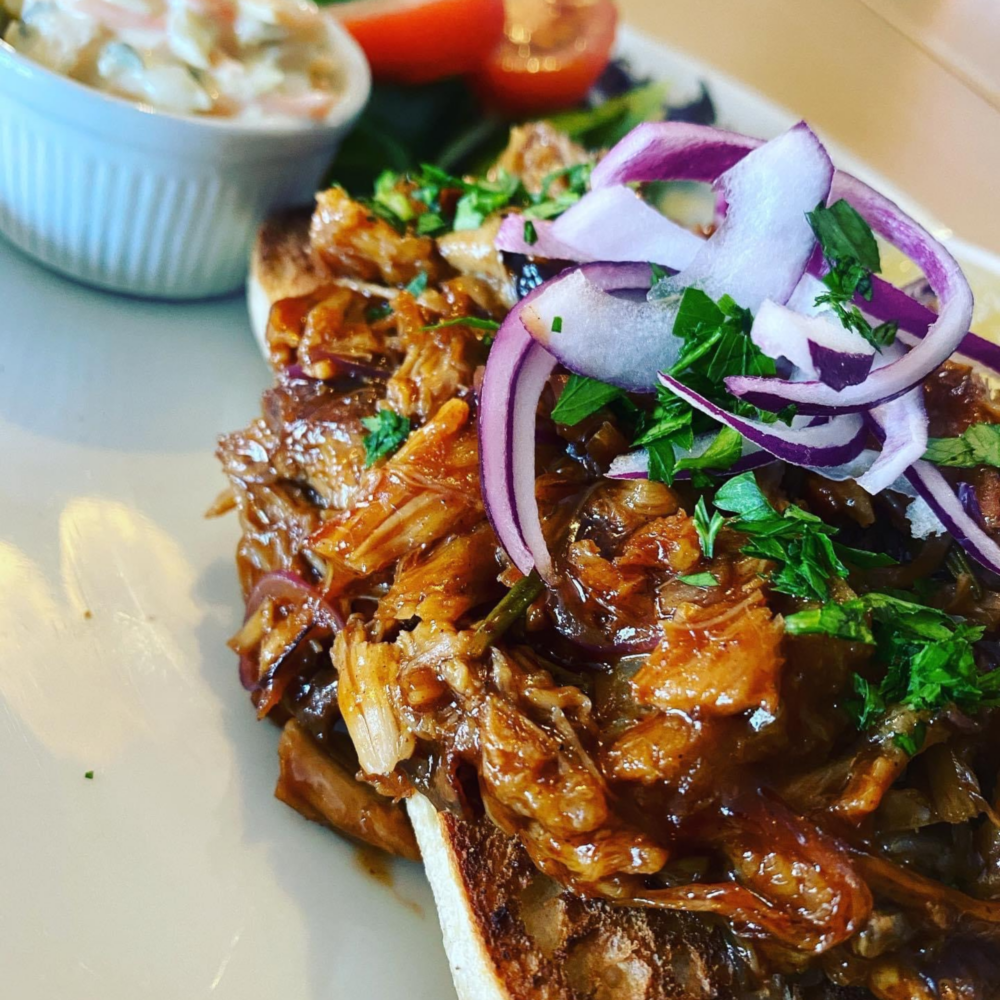 Pulled pork sandwich topped with red onion and herbs, with a side dish of coleslaw and fresh vegetables on the plate. - Home Instead