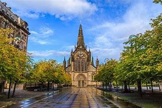 A majestic church with a tall spire, flanked by trees and wet pavement, under a partly cloudy blue sky. - Home Instead