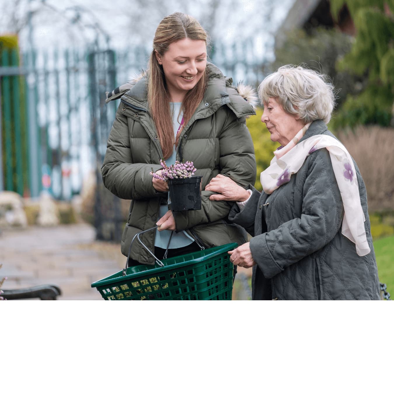 A young woman helps an older woman who is holding her arm. The older woman holds a garden basket and the younger woman a potted plant. - Home Instead
