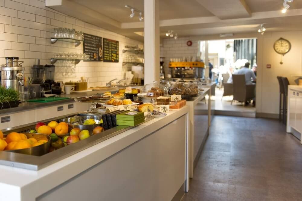 A bright, modern café interior with a counter displaying fruits, pastries, and desserts, with seating visible in the background. - Home Instead