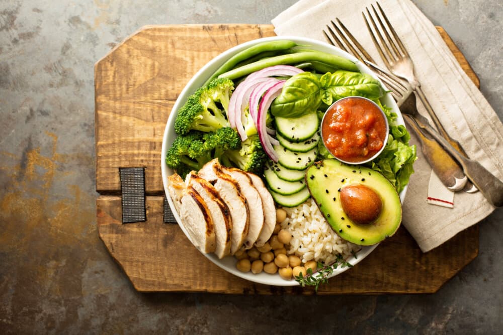 A bowl of sliced chicken, avocado, vegetables, rice, and chickpeas with a small cup of red sauce, on a wooden board. - Home Instead