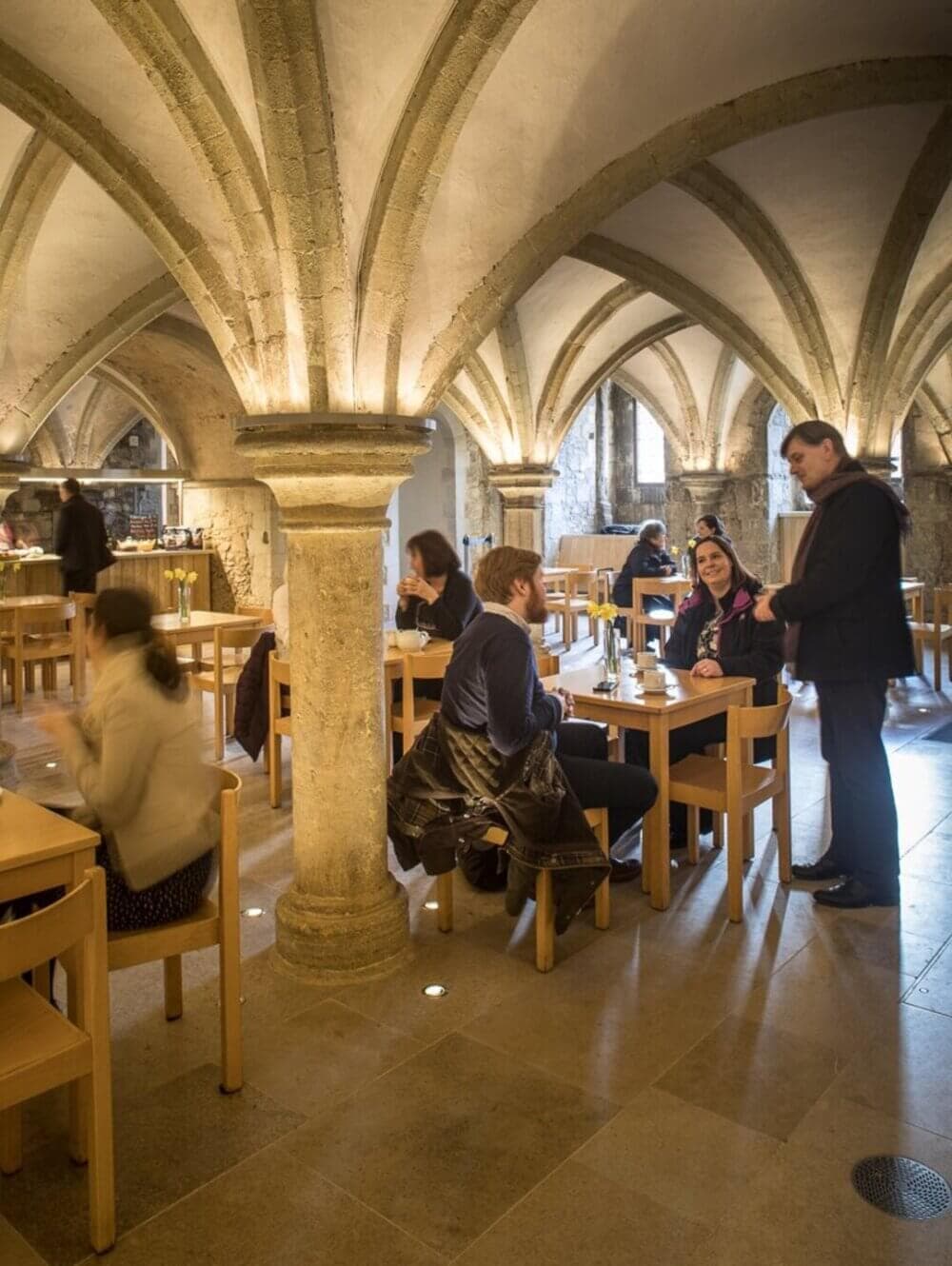 People dining and conversing at wooden tables in a vaulted, stone-arched room with soft lighting. - Home Instead
