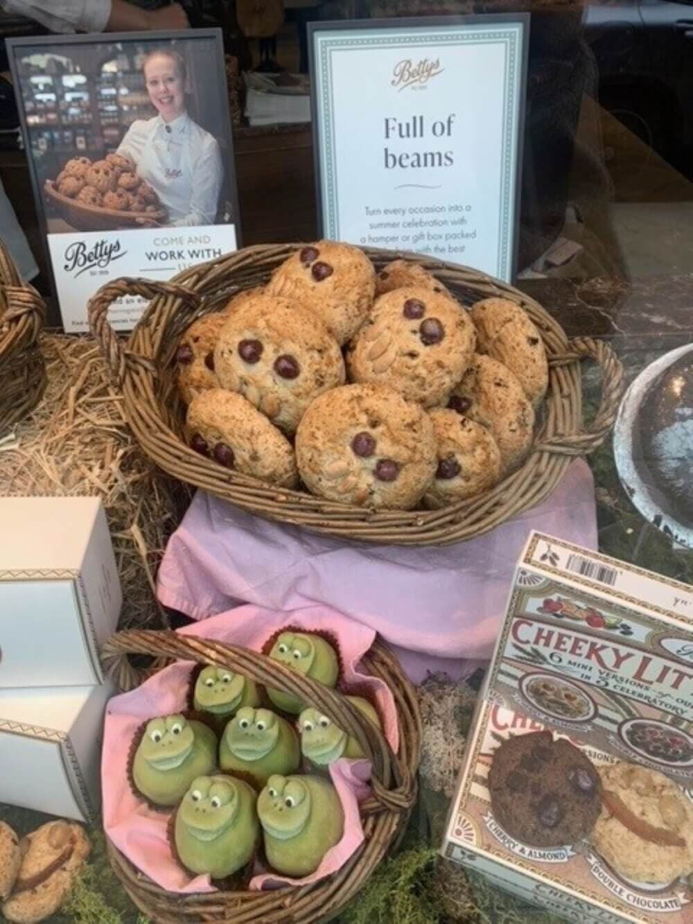 A bakery display with baskets of cookies, a sign "Full of beans," and frog-shaped desserts on a pink cloth. - Home Instead