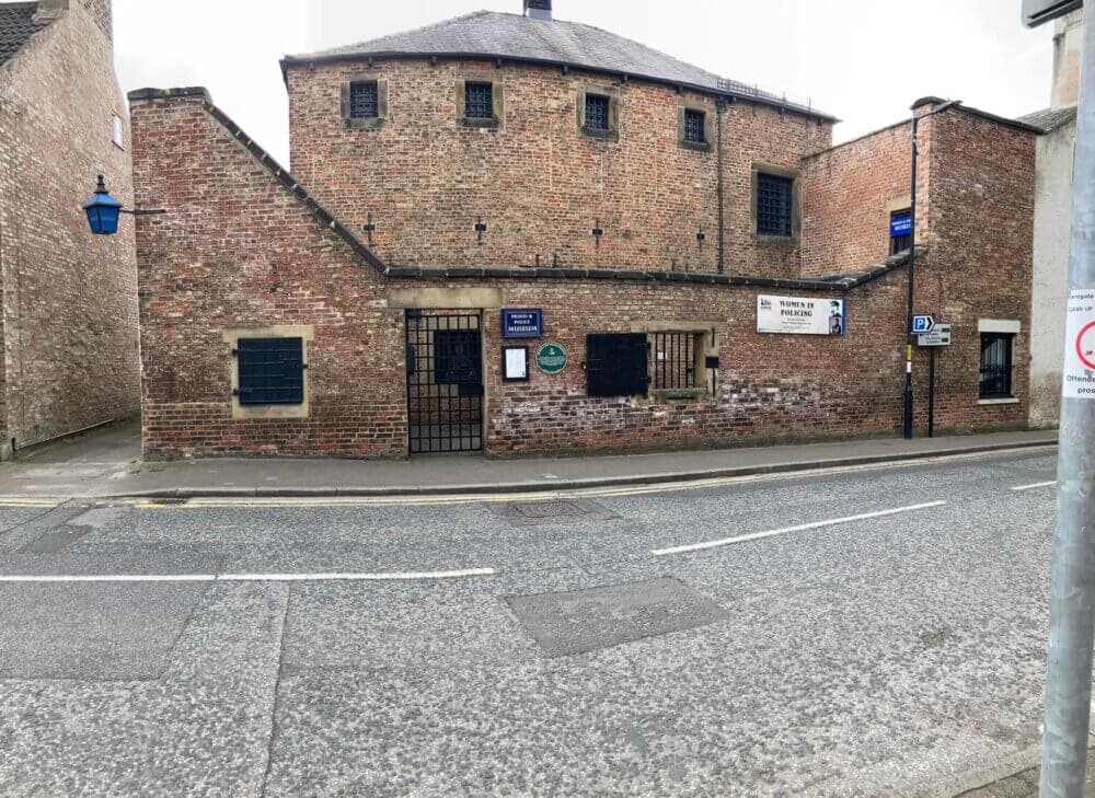 A historic brick building with barred windows and various plaques on the wall, seen from across a quiet street. - Home Instead