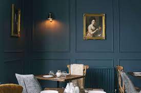Cozy dining area with dark blue walls, framed portrait paintings, wooden tables, cushioned chairs, and soft lighting. - Home Instead