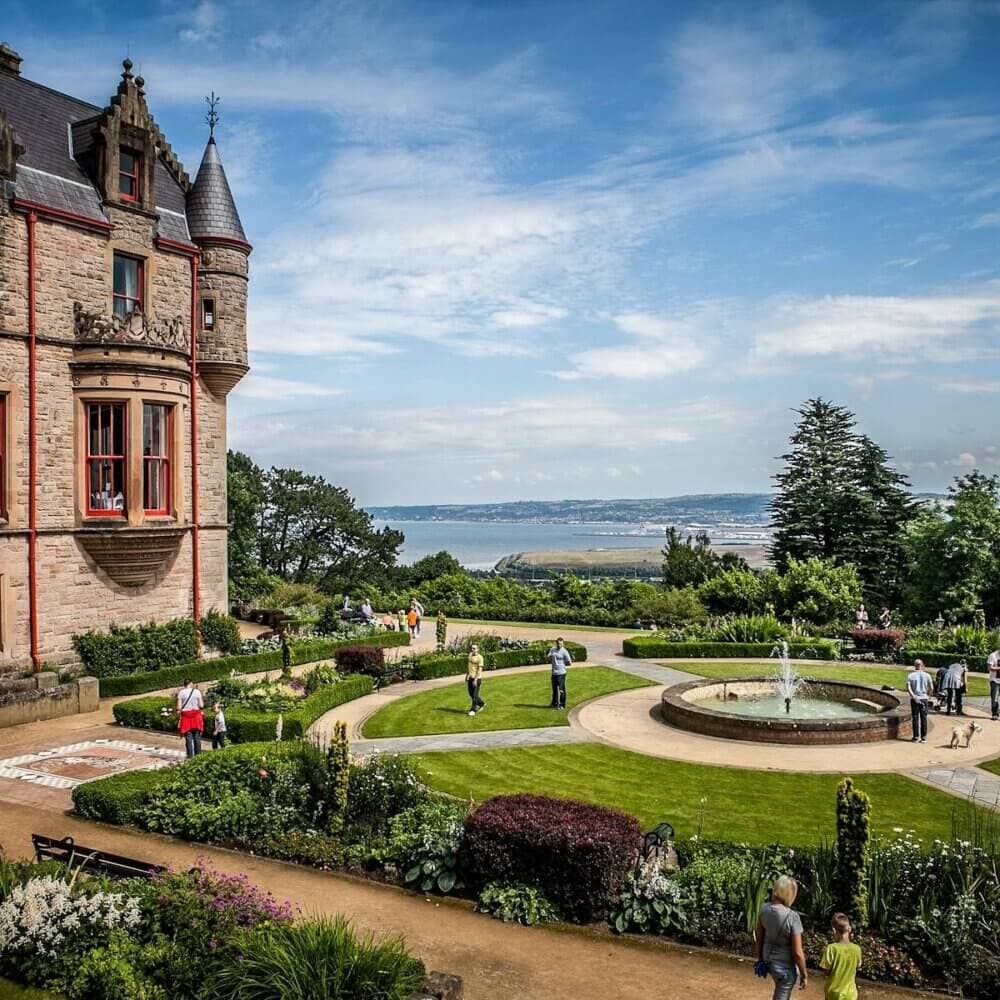 A scenic view of a castle with gardens and a fountain, set against a backdrop of hills and a large body of water. - Home Instead