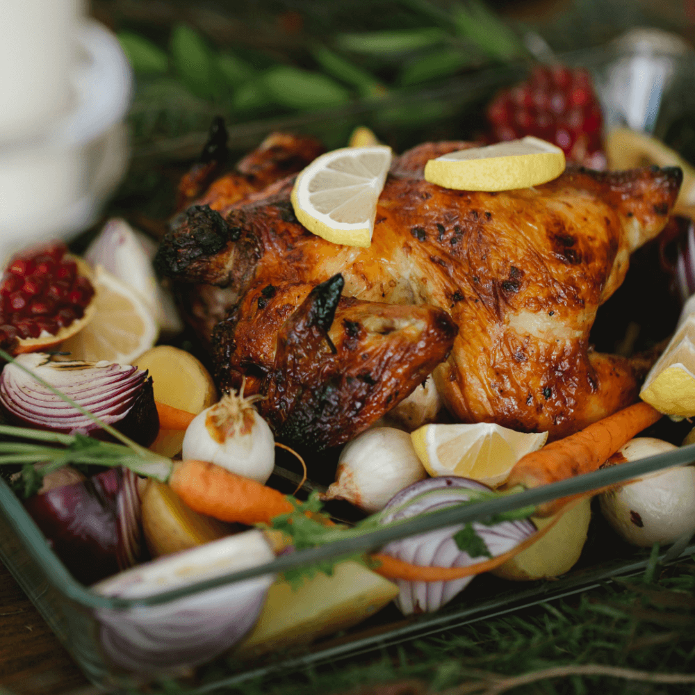 Roasted chicken garnished with lemon slices, surrounded by carrots, onions, garlic, and pomegranate in a glass dish. - Home Instead