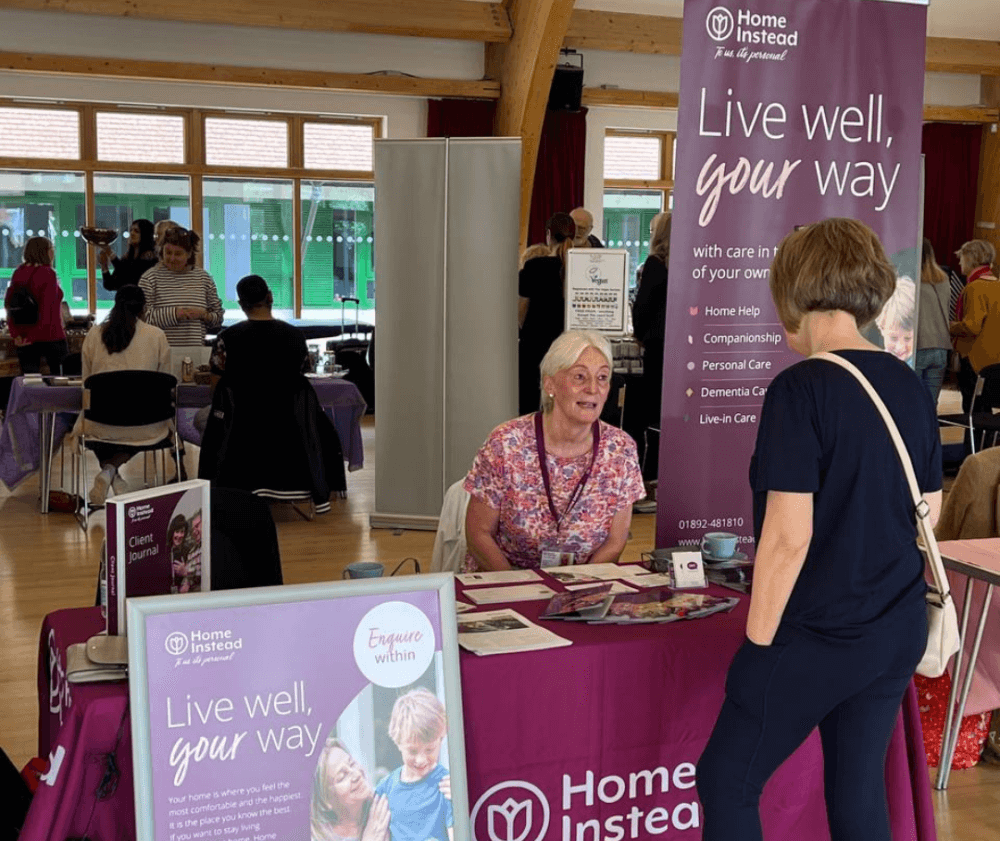 People interacting at an information booth for Home Instead with a "Live well, your way" banner in a community center. - Home Instead