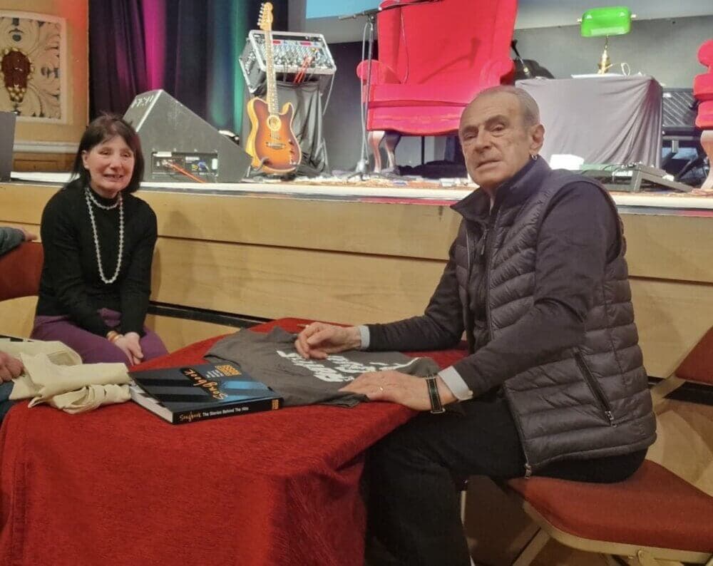Two people at a table, one of them sitting and signing a t-shirt. Musical instruments on stage in the background. - Home Instead