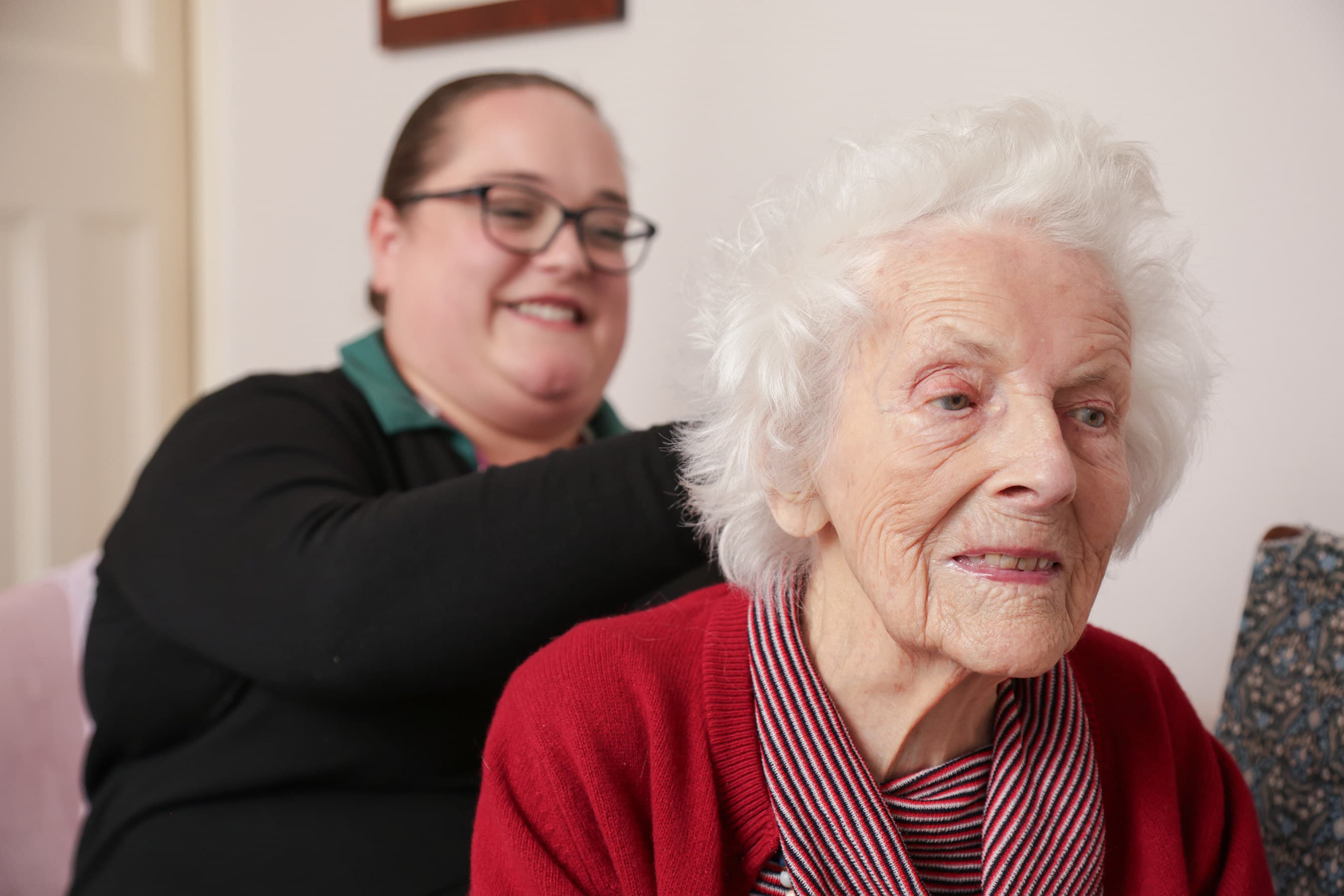 Older woman in a red sweater sits as a younger woman smiles behind her, possibly helping with her hair. - Home Instead