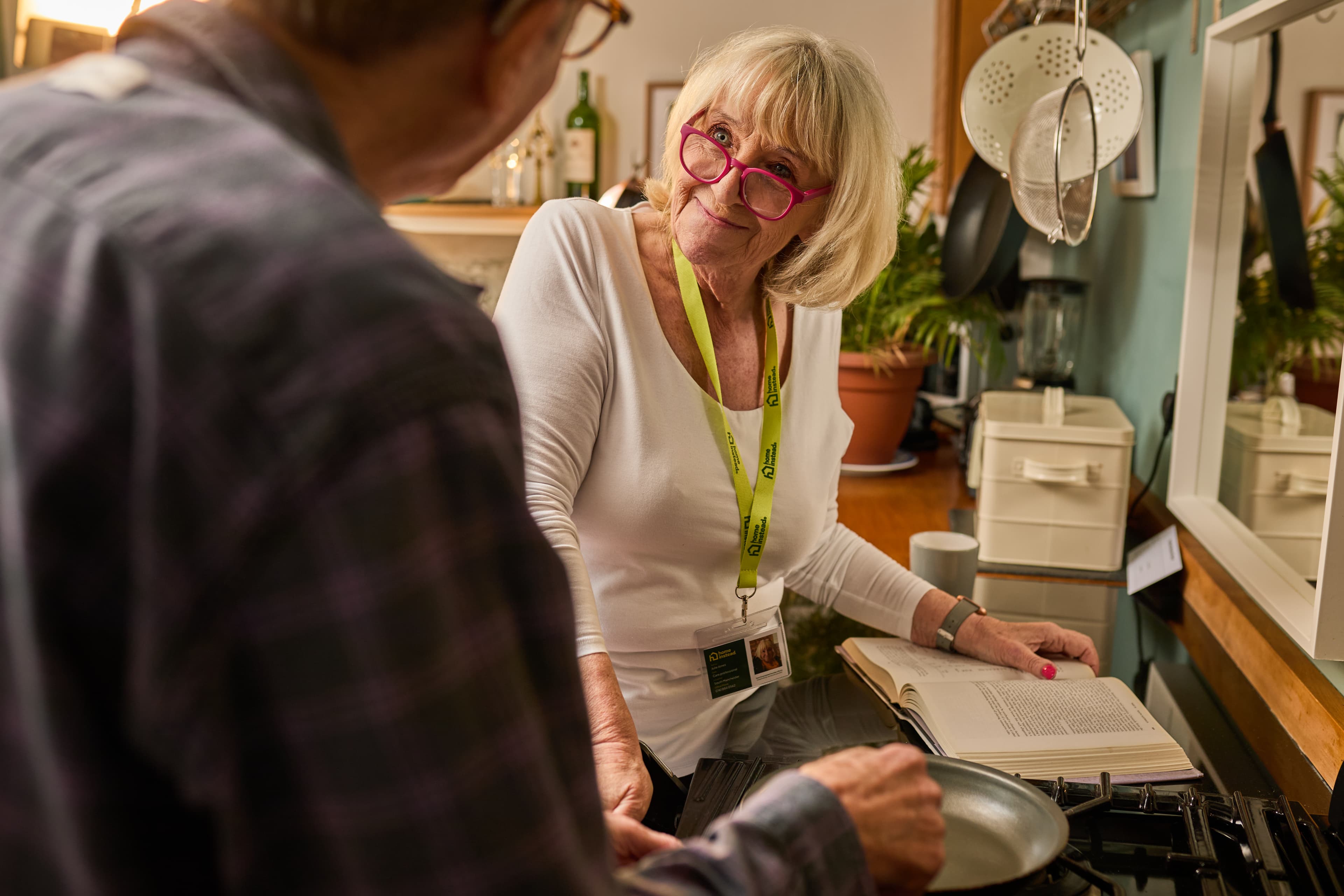Older woman with pink glasses talks to a man in a kitchen, holding a pan with an open cookbook nearby. - Home Instead