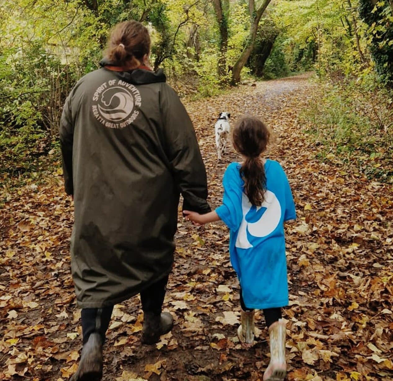 An adult and child hold hands walking a leaf-covered forest path, following a white dog ahead. - Home Instead