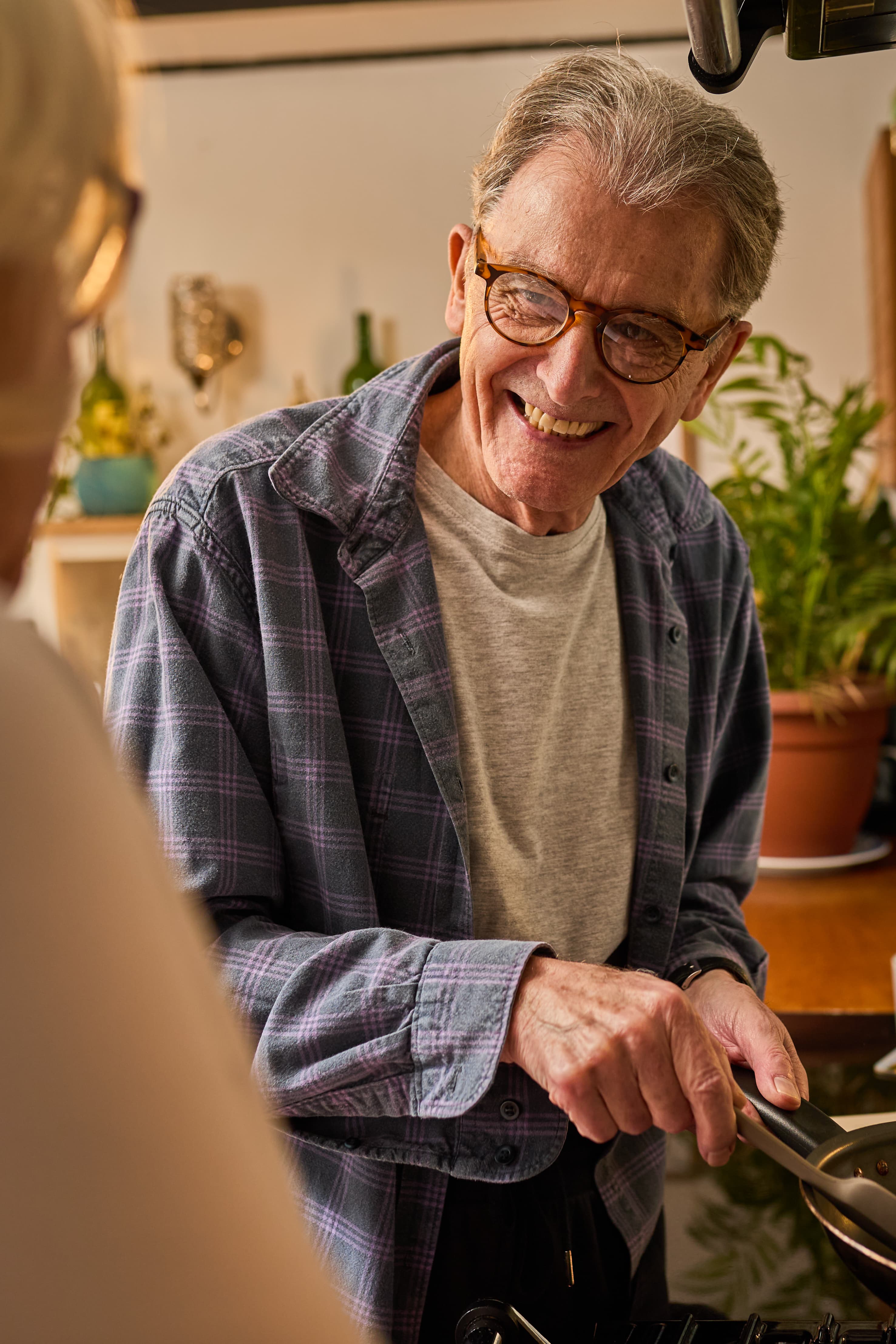 An older man in glasses smiles whilst cooking at home, holding a frying pan and talking to someone nearby. - Home Instead