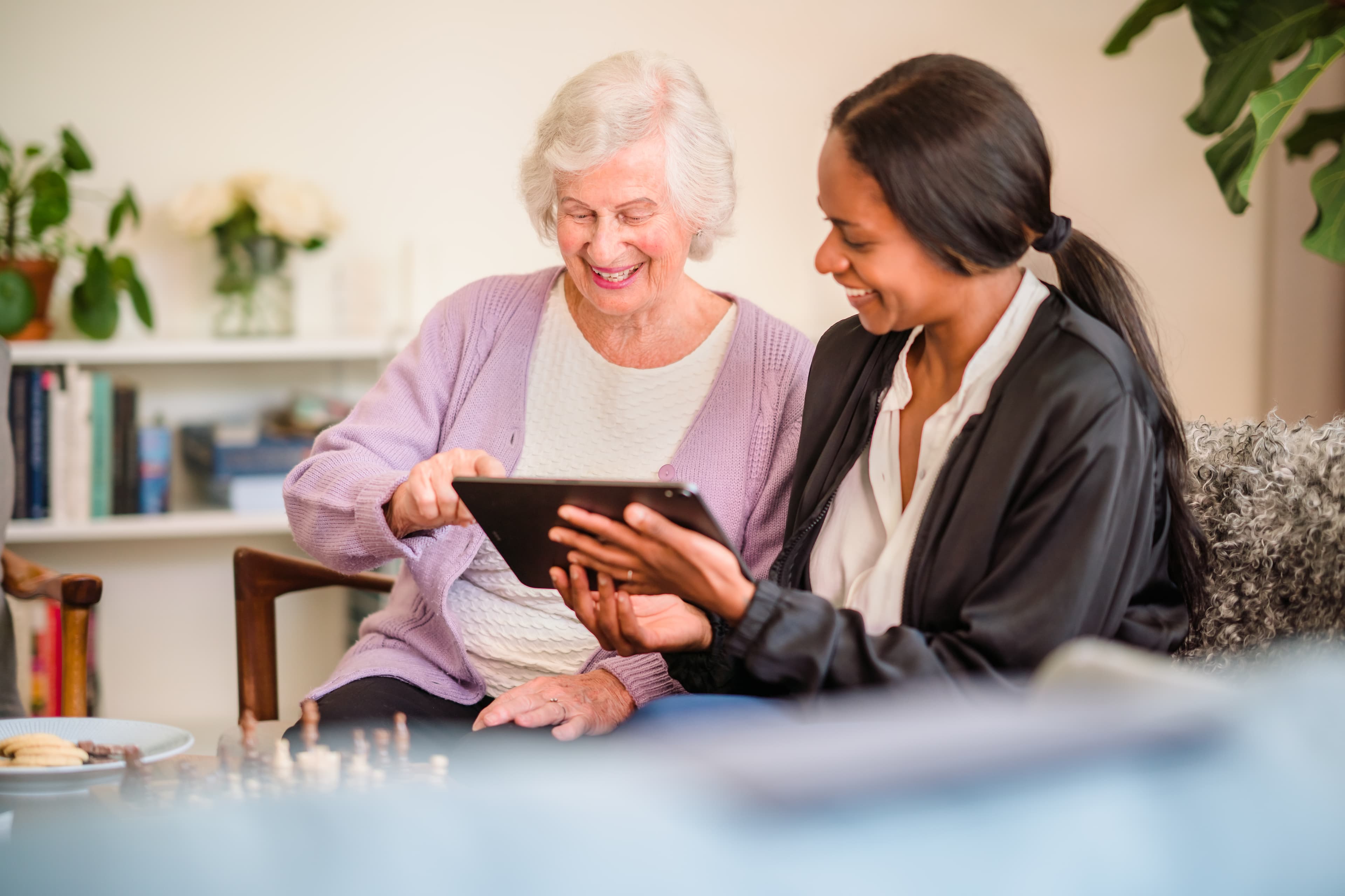 An elderly woman and a younger woman sit together on a couch, smiling and looking at a tablet. - Home Instead
