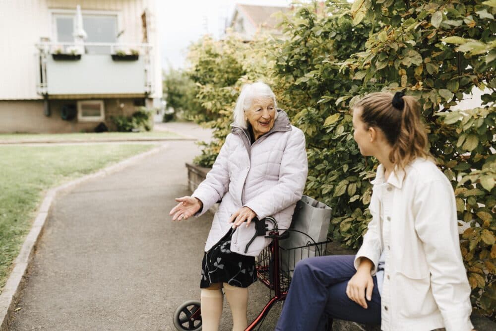 An elderly woman on a walker chats with a younger woman on a garden path. - Home Instead