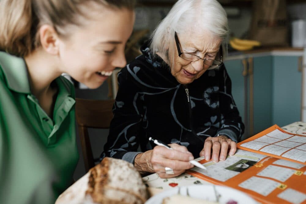 Elderly woman writing on a calendar while a young woman smiles beside her at a table. - Home Instead