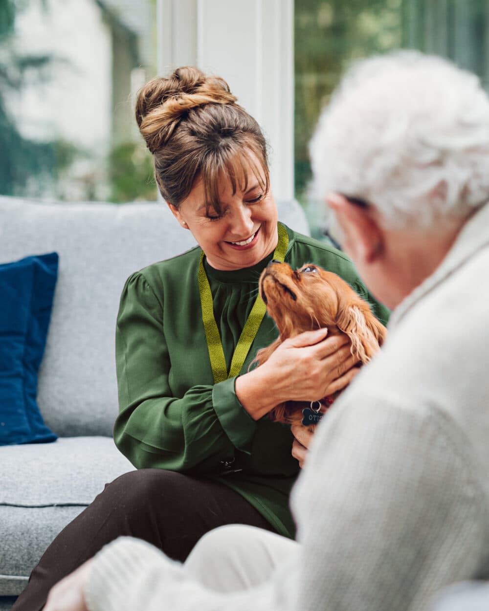 A woman smiling and petting a small brown dog while sitting next to an elderly man indoors. - Home Instead