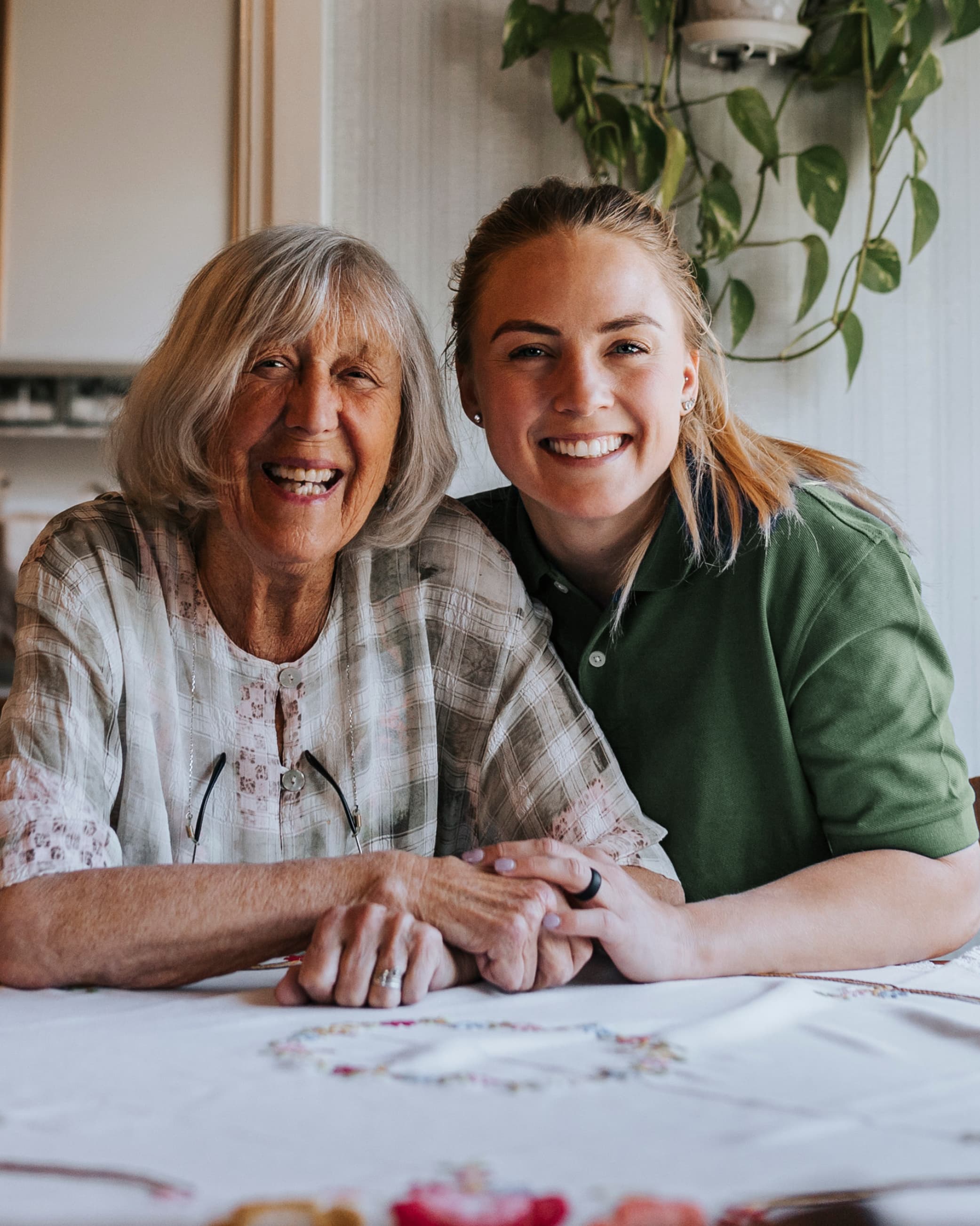 Smiling elderly woman and young woman sit together at a table, holding hands in a cosy home setting. - Home Instead