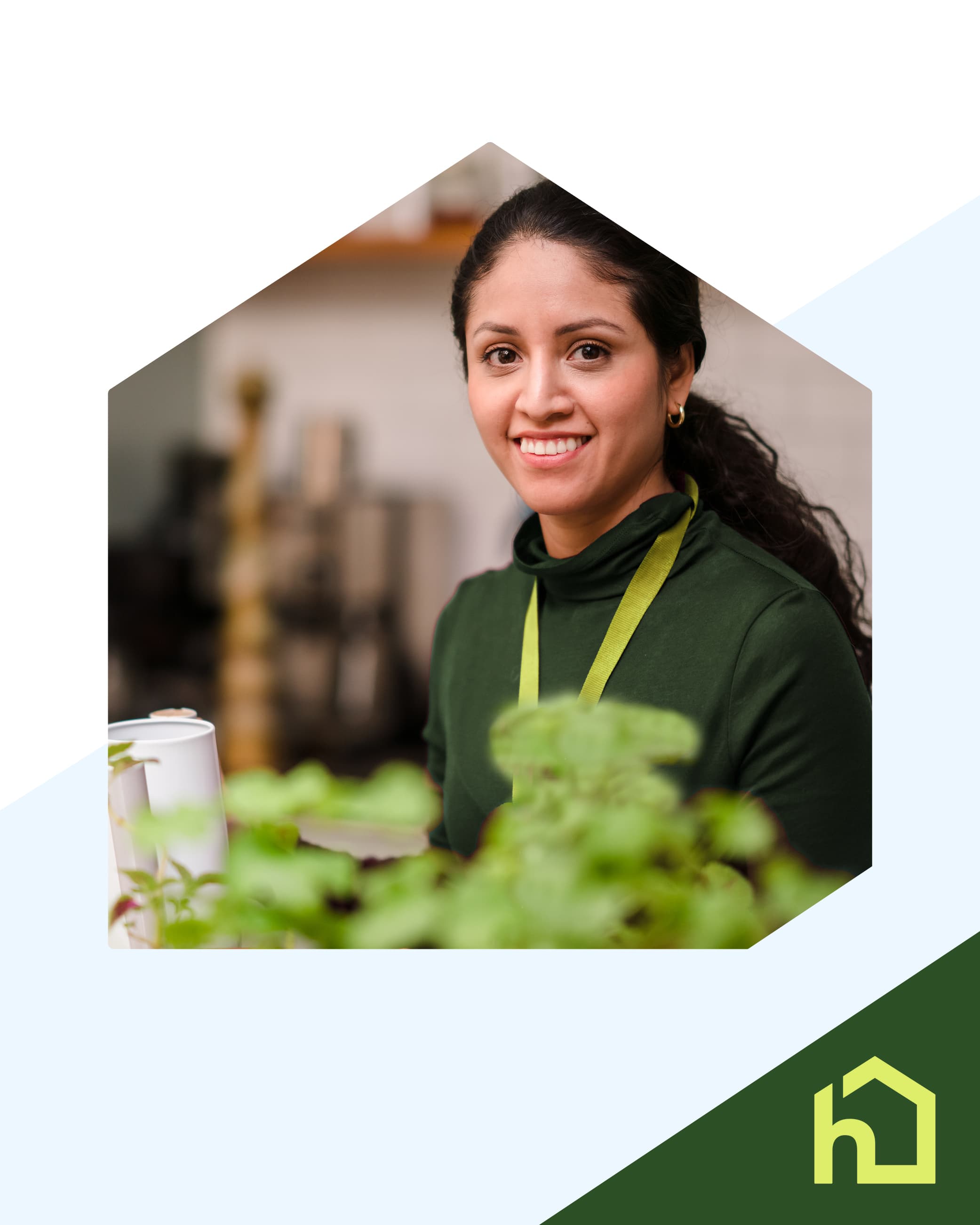 A smiling woman in a green top and apron sits behind potted plants in a bright room. - Home Instead