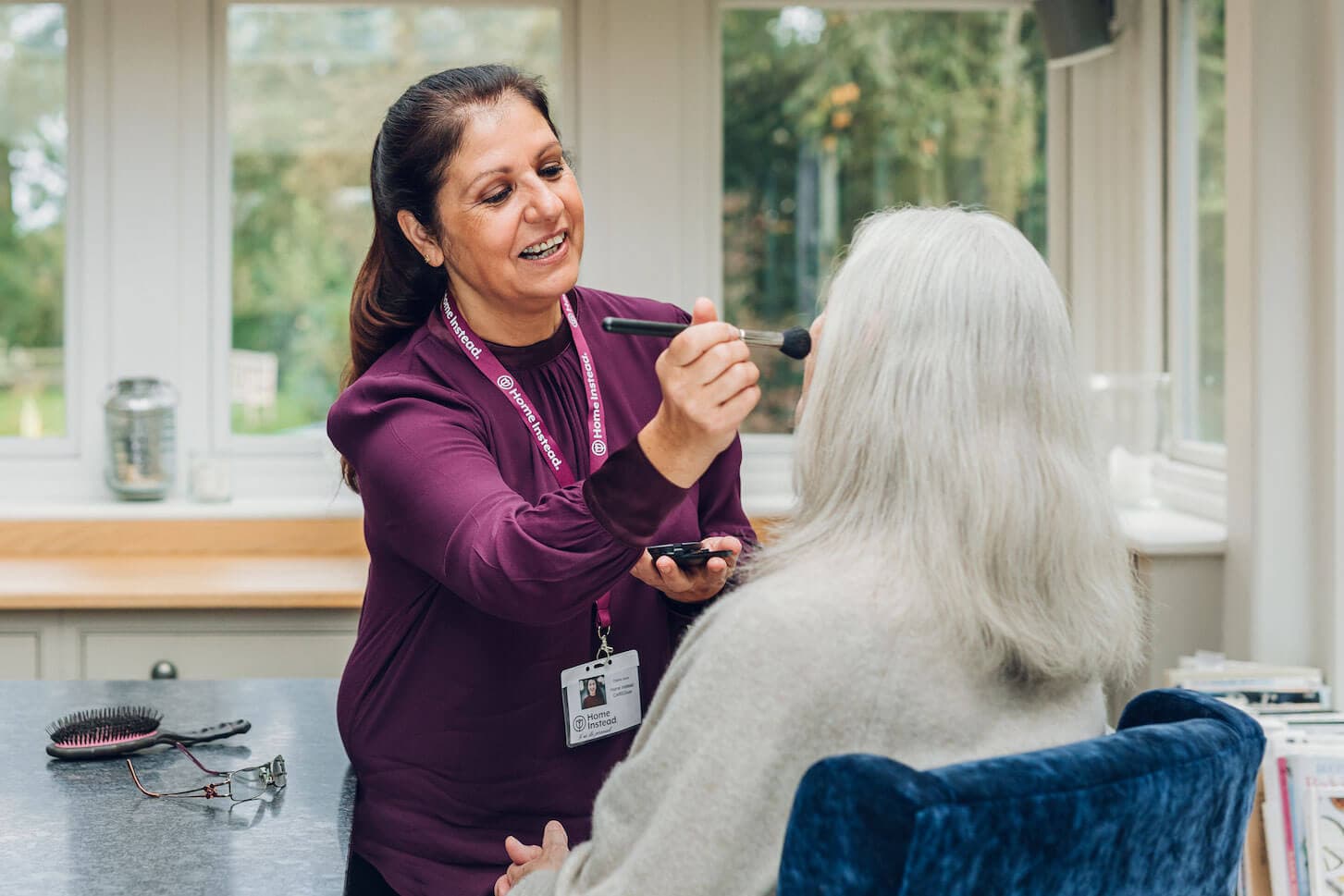 A caregiver applies makeup to an elderly woman seated in a chair, in a bright room with large windows. - Home Instead
