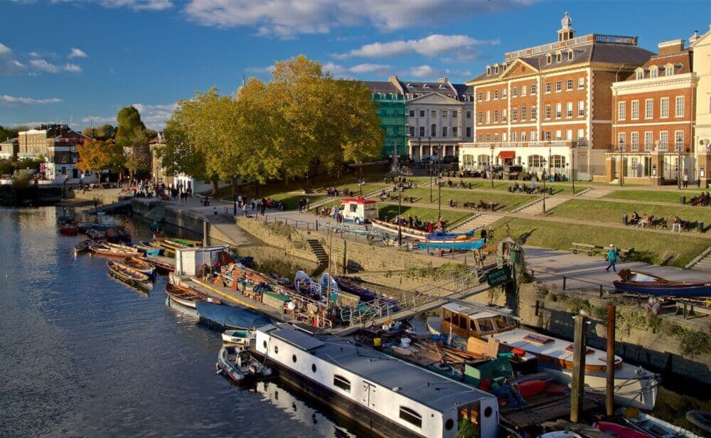 People relaxing on grassy steps by the riverside, with boats docked in the water and buildings in the background. - Home Instead