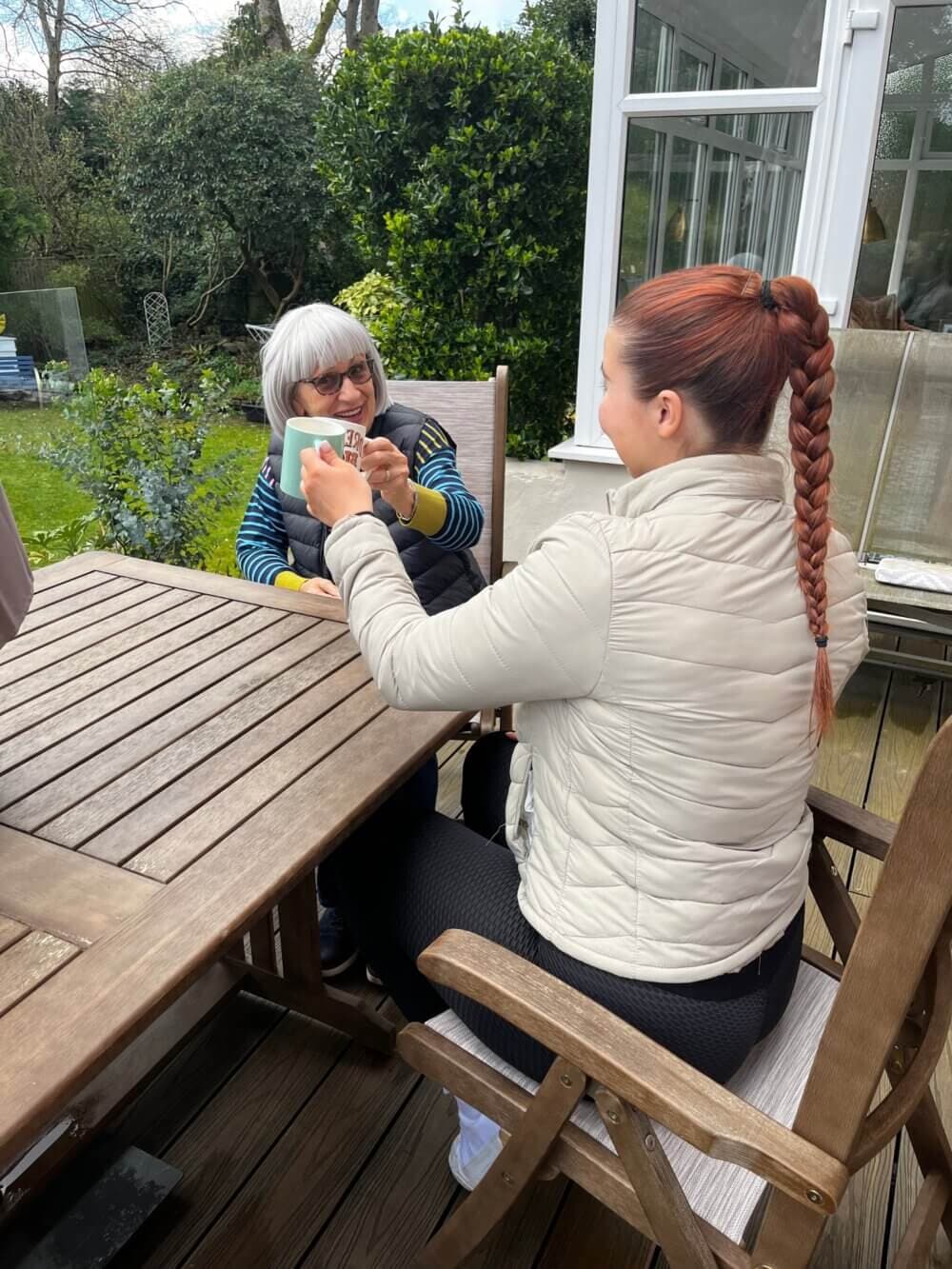 Two women sitting at an outdoor table, one with gray hair and glasses, the other with a long braid, sharing a laugh. - Home Instead