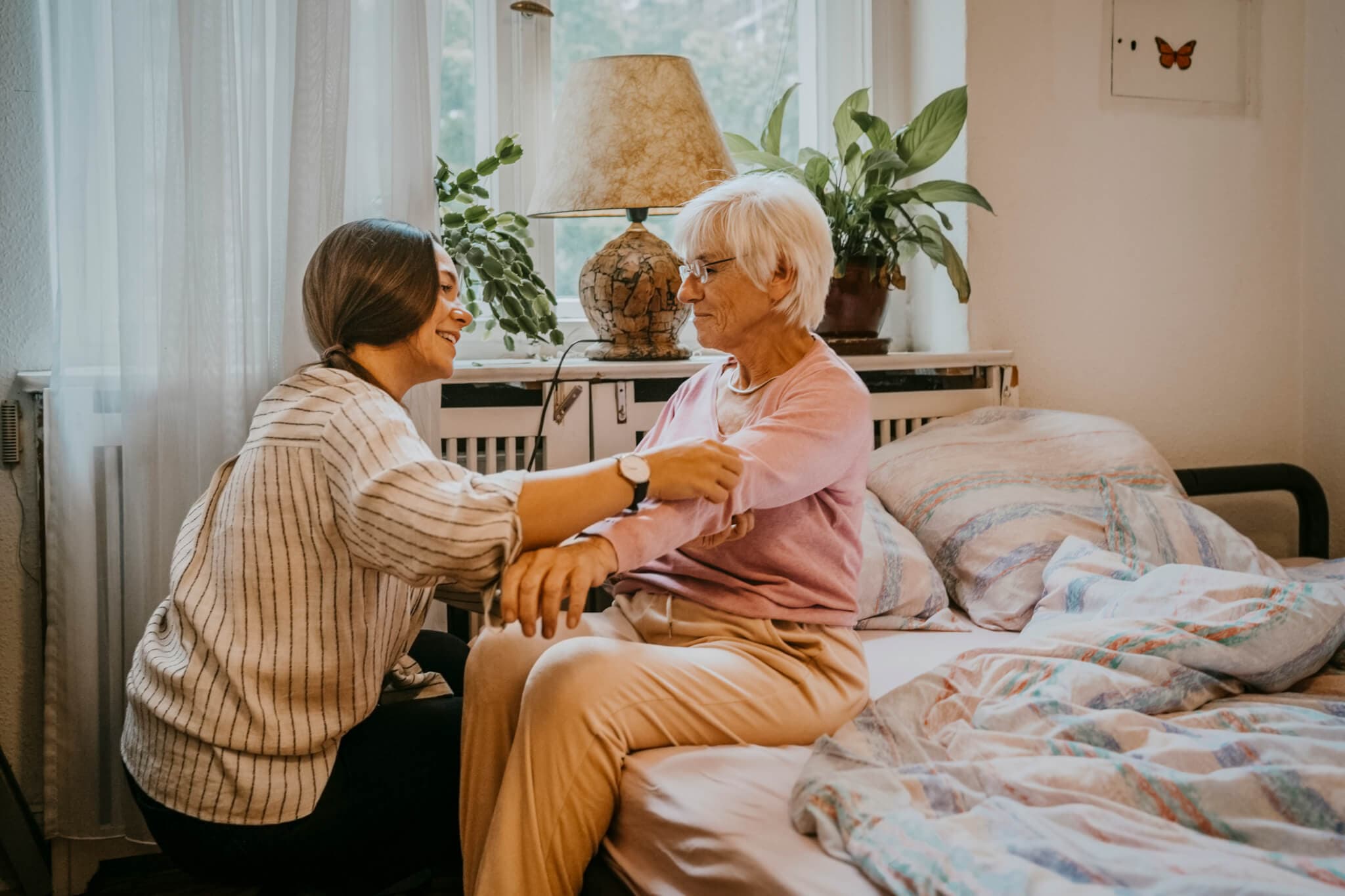 A young woman helps an elderly woman get up from her bed in a cozy, plant-filled room. - Home Instead