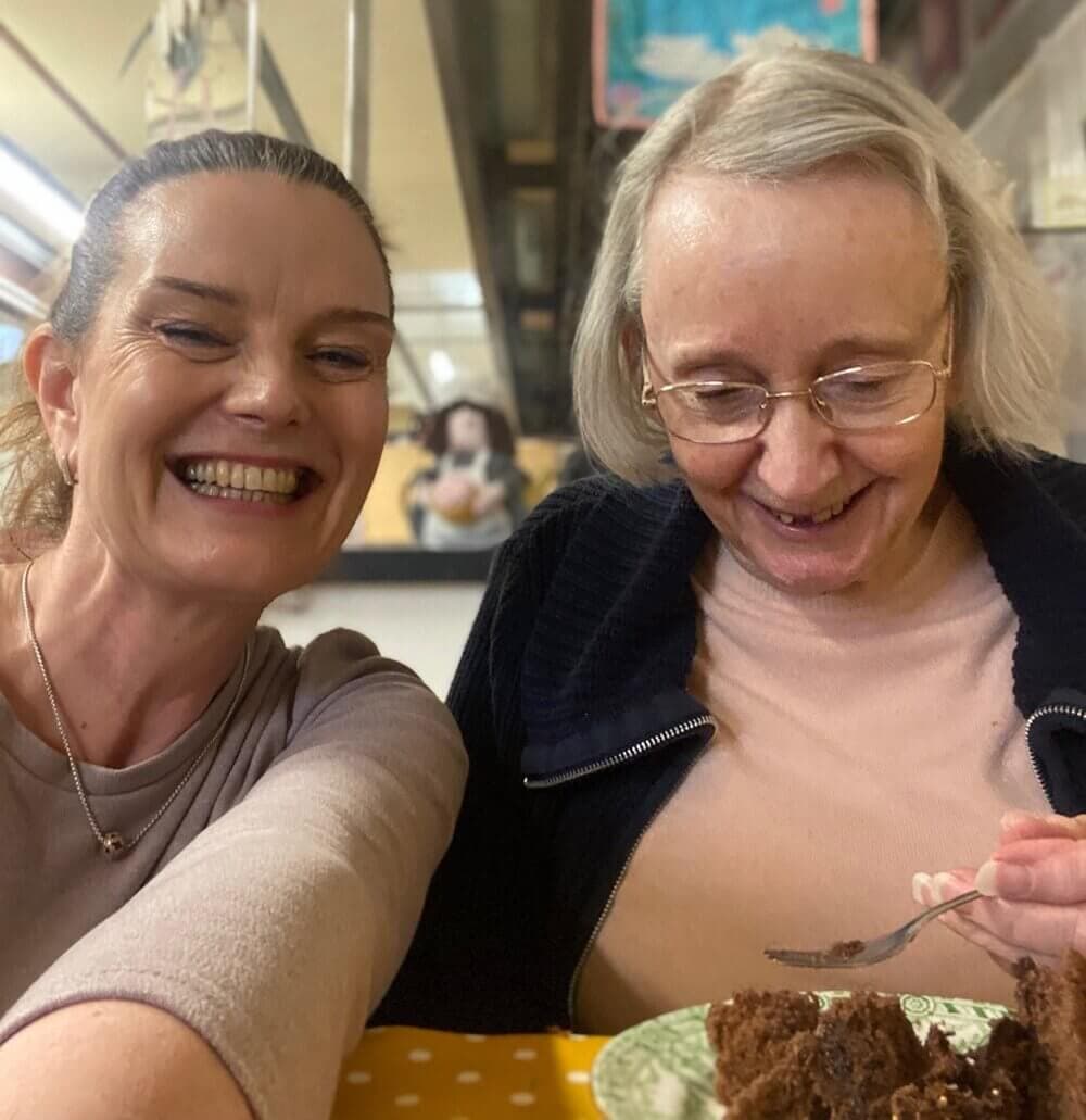 Two women smiling; one holding a fork and enjoying a piece of chocolate cake at a dining table. - Home Instead