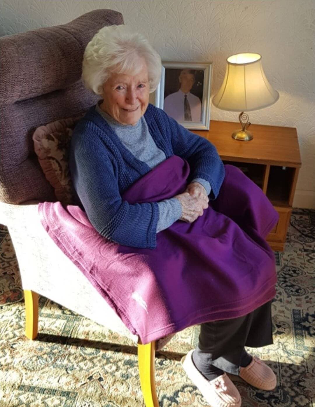 An elderly woman sits in a comfortable chair with a purple blanket, smiling, with a lamp and a framed photo nearby. - Home Instead