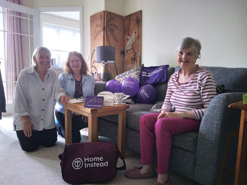 Three smiling women sit in a living room with Home Instead and Scope items, including bags, mugs, and balloons. - Home Instead