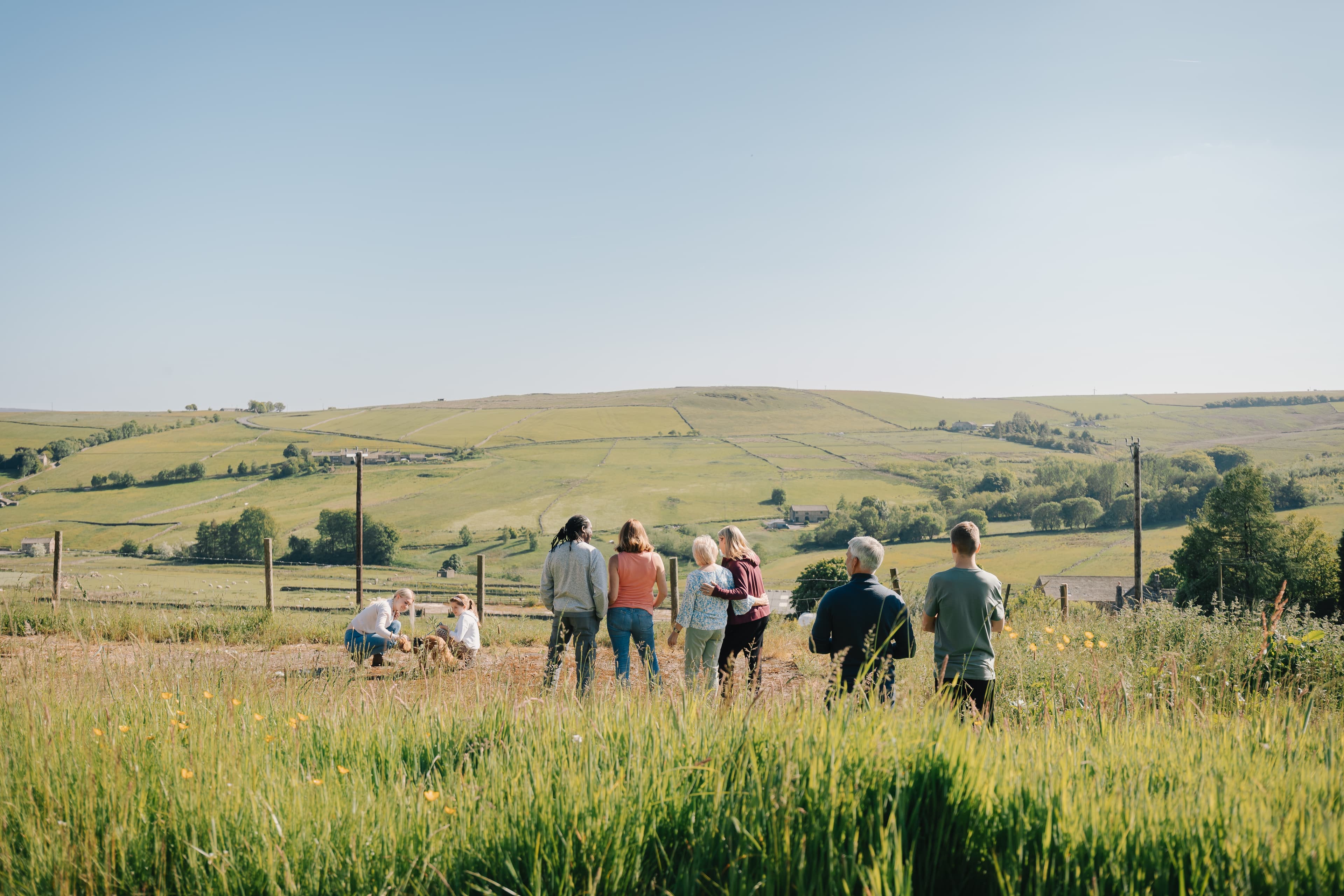 Group of people walking through a grassy field with a hilly landscape in the background under a clear blue sky. - Home Instead