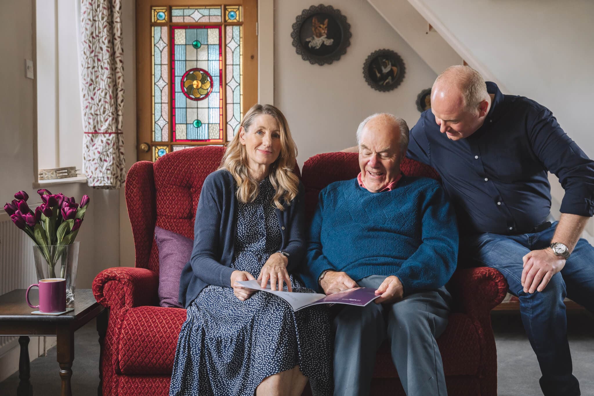 Three people smile while sitting on a red couch, looking at a book together in a cozy living room with a stained glass door. - Home Instead