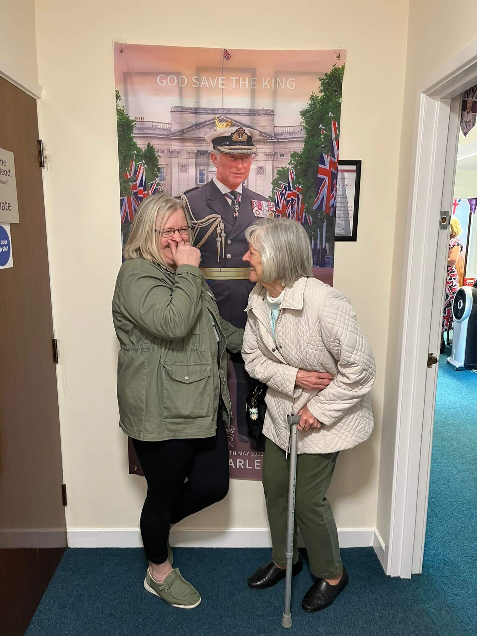 Two women, one elderly with a cane, stand laughing in front of a "God Save The King" poster. - Home Instead