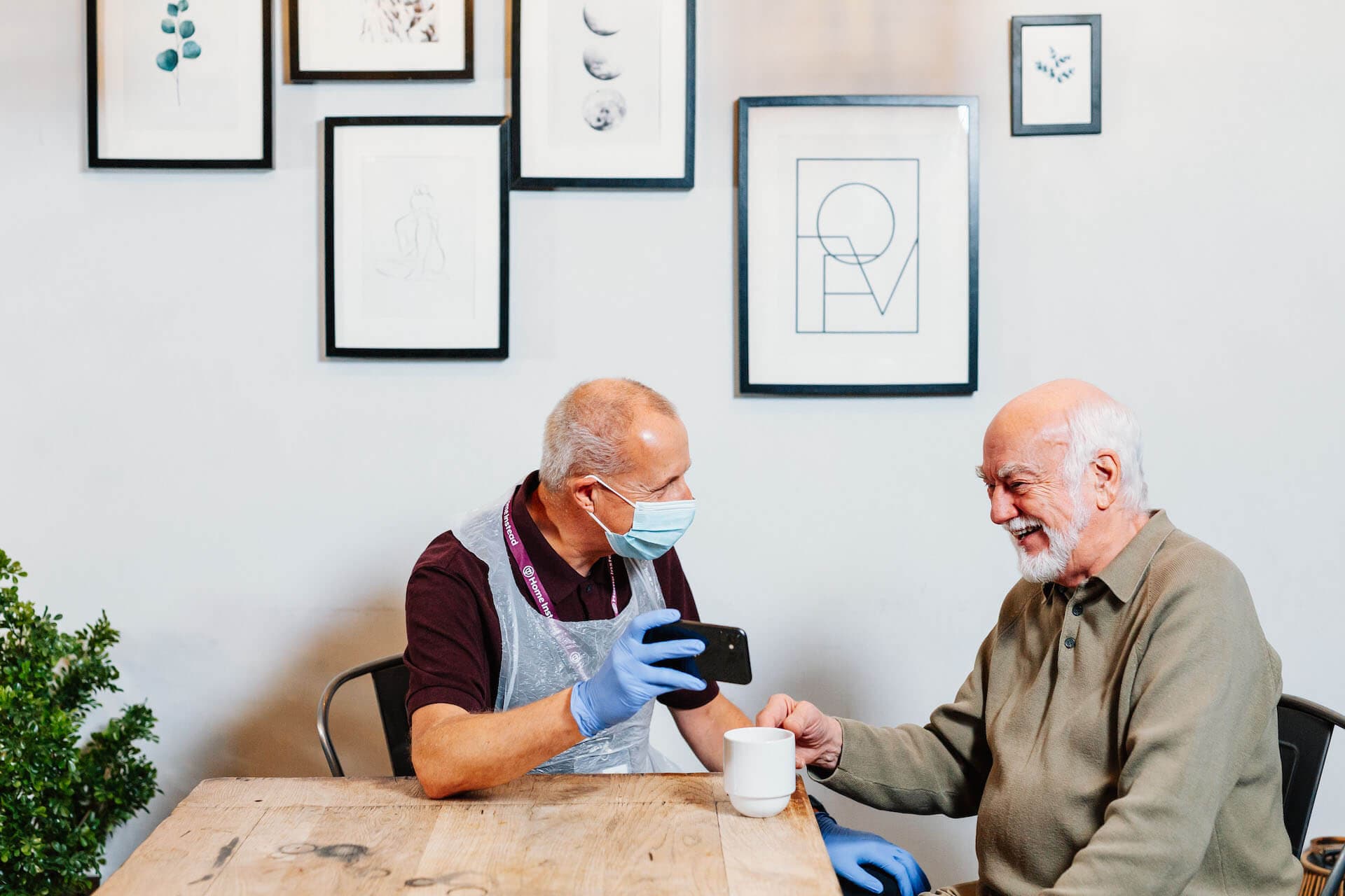 Two elderly men sitting at a wooden table, one showing the other a phone, both smiling and surrounded by wall art. - Home Instead