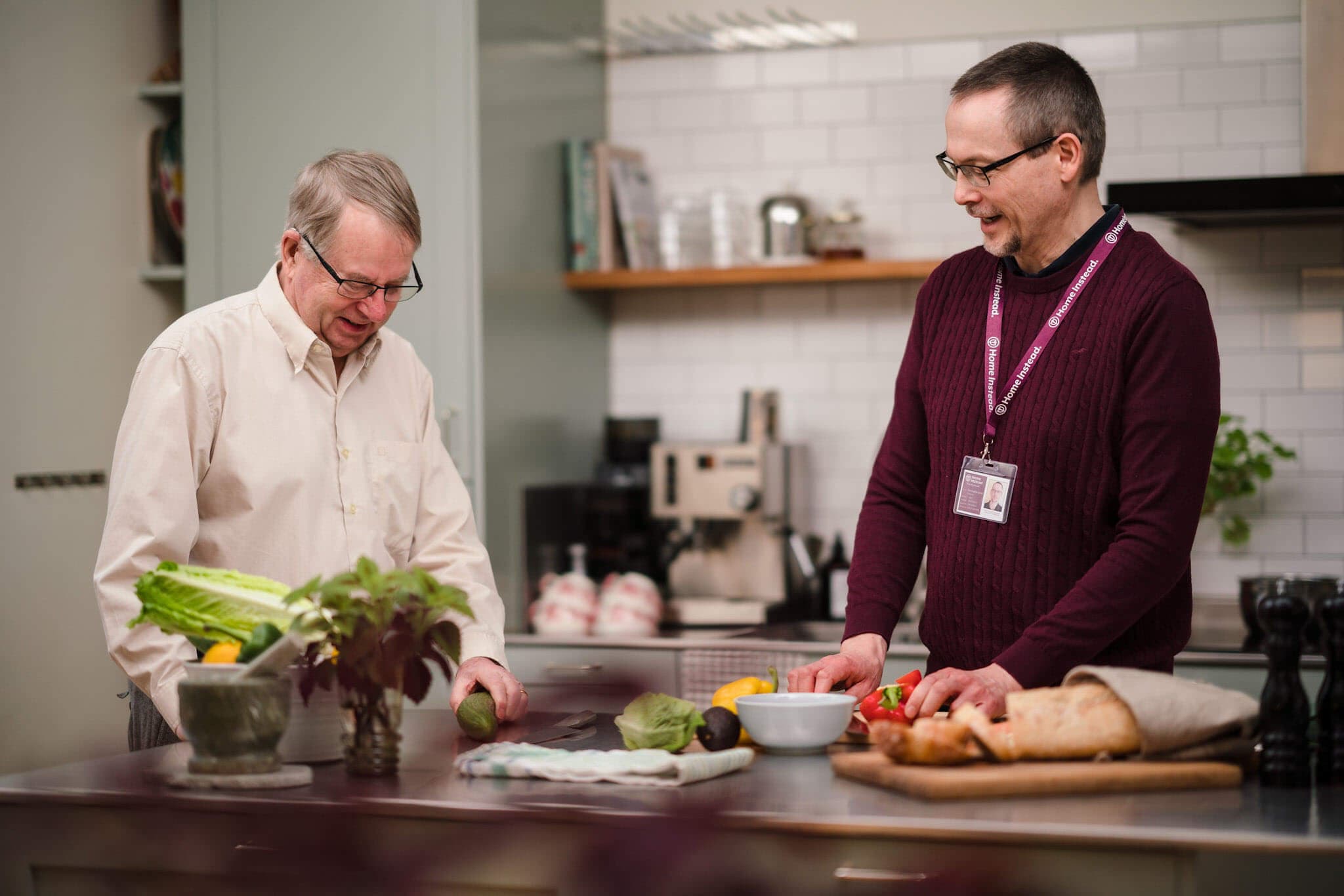 Two men preparing food together in a modern kitchen, surrounded by fresh vegetables and bread on the counter. - Home Instead