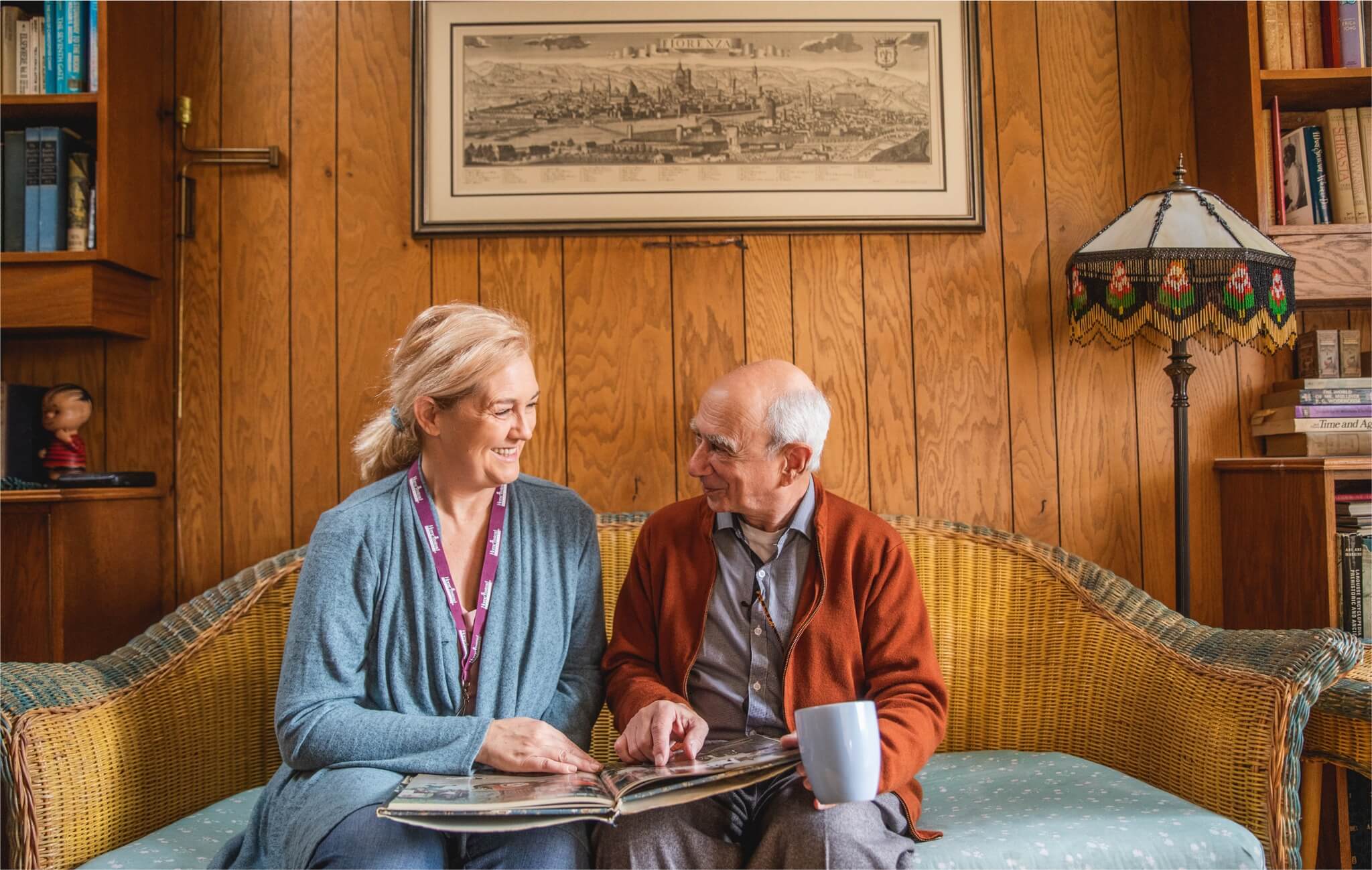 A woman and an elderly man sit on a couch, smiling, while looking at a photo album in a cozy, wood-paneled room. - Home Instead