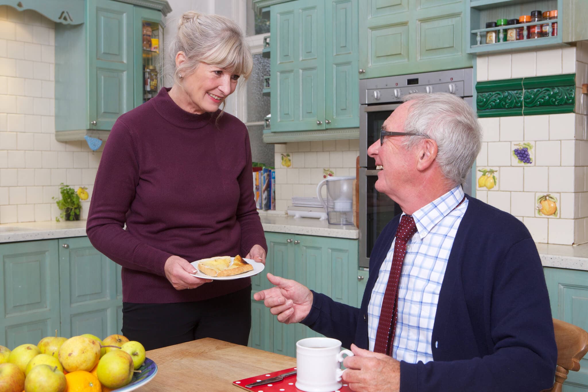 Woman serving food to seated man in a kitchen with green cabinets and a basket of apples on the table. - Home Instead