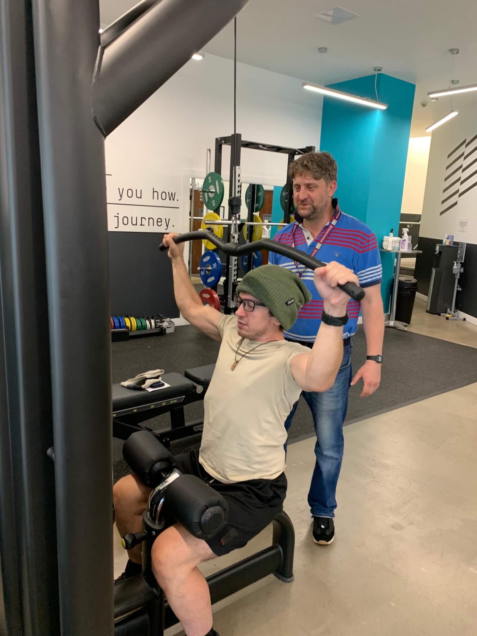 Person using a lat pulldown machine in a gym, assisted by another individual standing behind them. - Home Instead