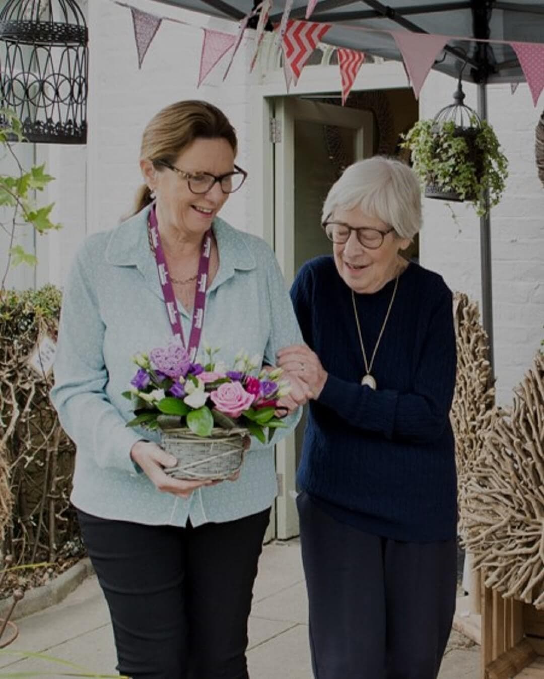 Two women, one holding a flower basket, walk together under a canopy with hanging decorations. - Home Instead