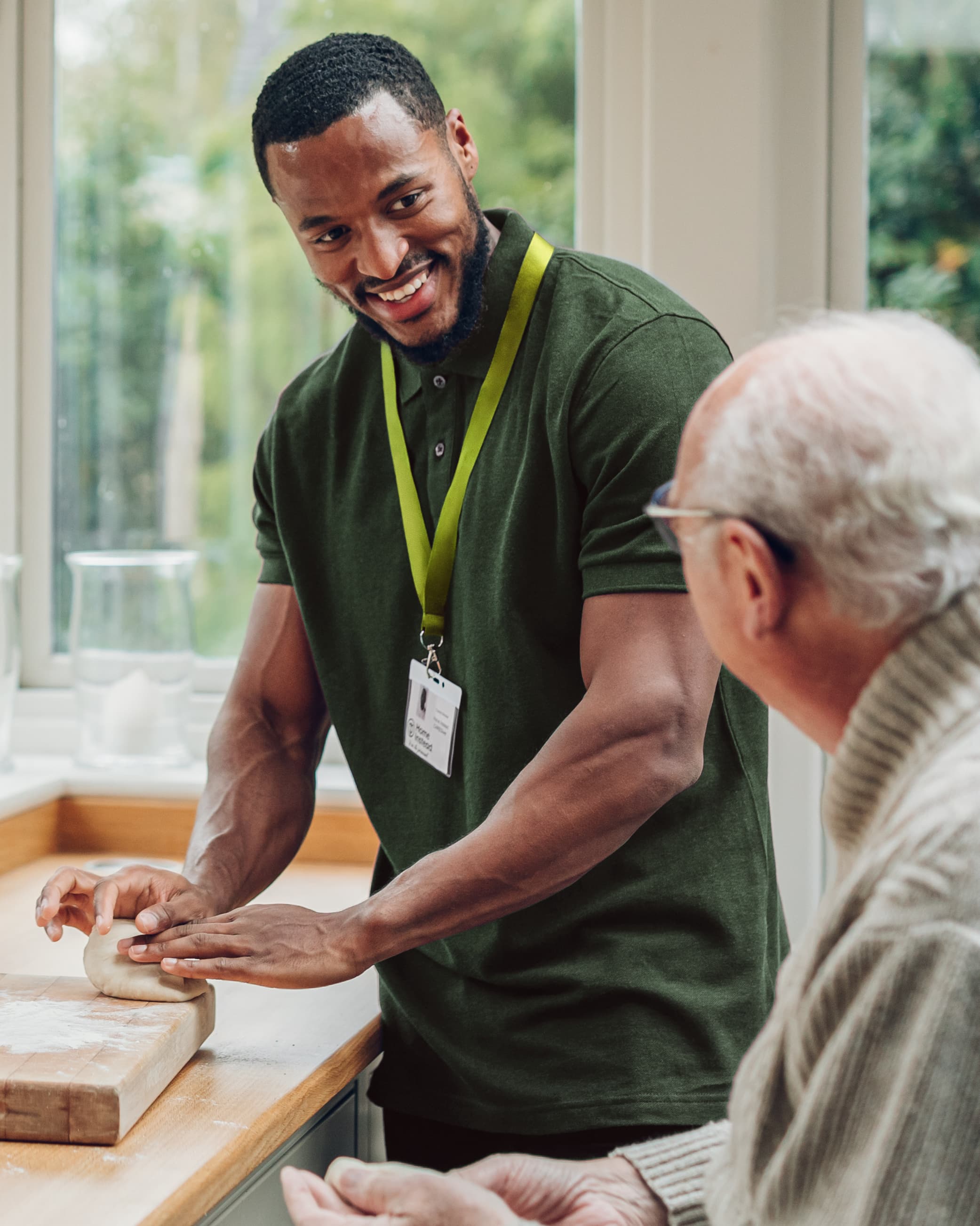 A caregiver helps an elderly man knead dough, both smiling in a bright kitchen. - Home Instead